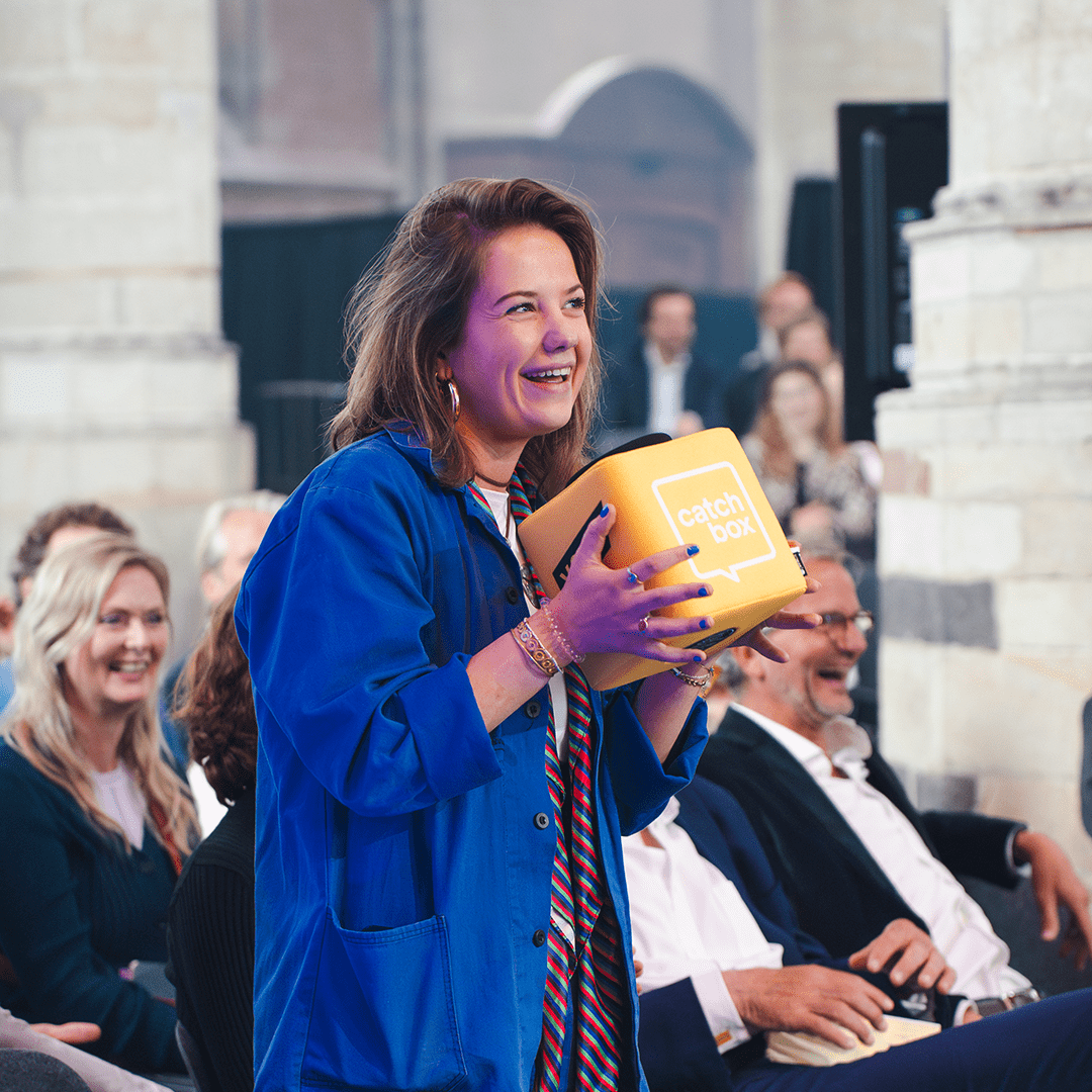 Conference attendee holding a customized yellow Catchbox Cube microphone during a Q&A session, with an engaged audience in the background.