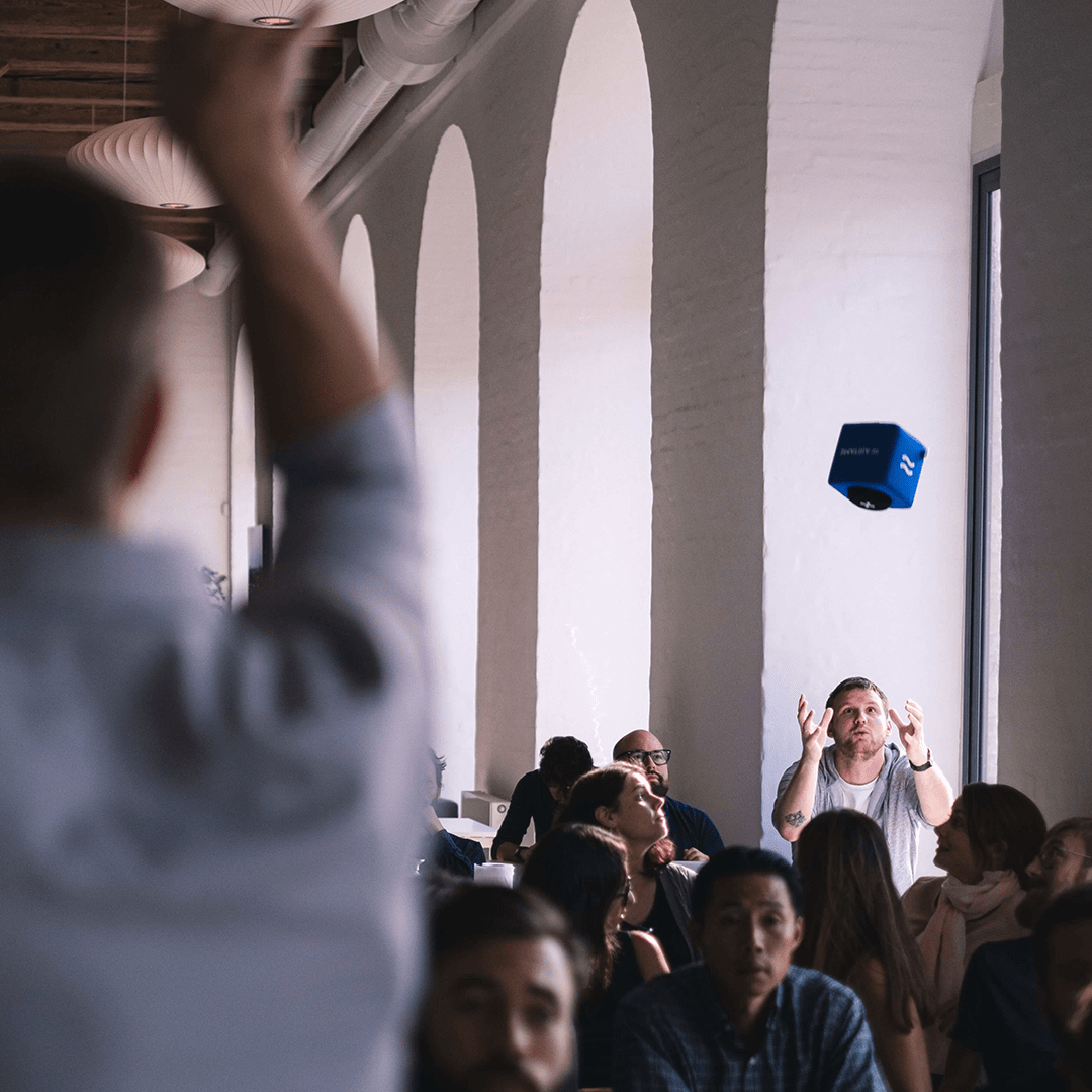 Audience member catching a blue Catchbox Cube microphone being tossed across a well-lit room with arched windows, engaging in an interactive session.