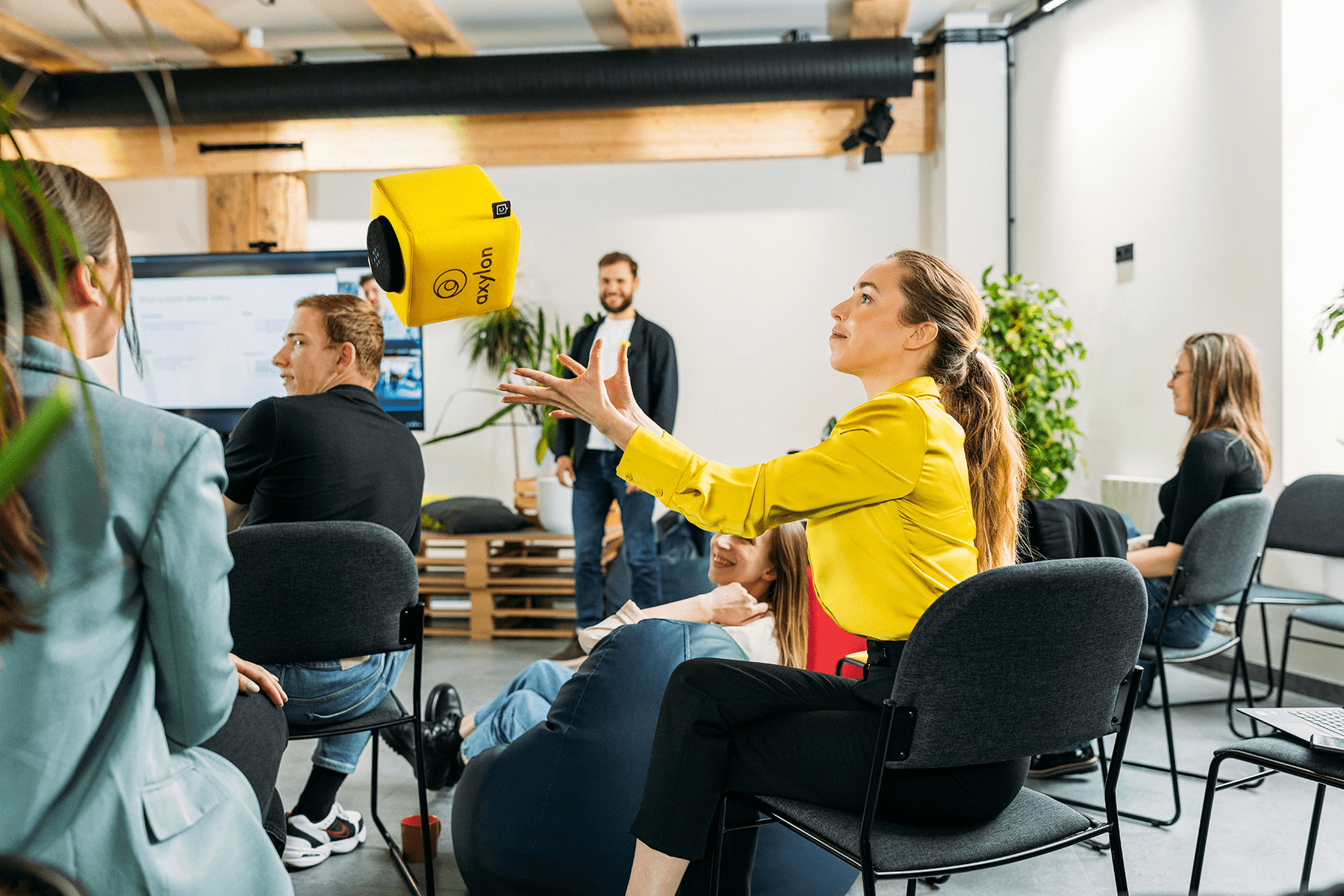 A woman in a yellow shirt catching a yellow Catchbox Cube microphone during a collaborative session, with other participants seated and engaging in the background.