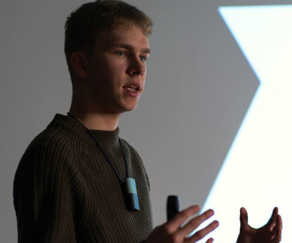 Presenter wearing a Catchbox Clip microphone, gesturing while delivering a speech, emphasizing how using microphones can prevent voice strain and ensure clear, impactful presentations at events.