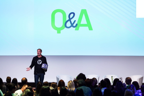 Presenter on stage holding a Catchbox Cube microphone with a Q&A sign displayed on the screen behind him, engaging the audience and highlighting the importance of avoiding common audio mistakes to keep events interactive and engaging.