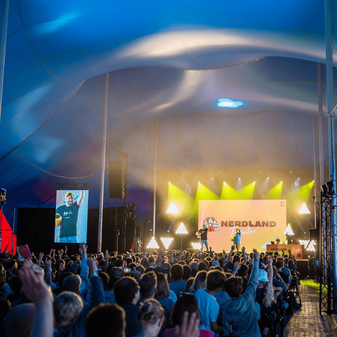 Person on stage using a Catchbox Cube microphone, engaging a large audience at the Nerdland event under a tent, with vibrant lighting and an enthusiastic crowd.