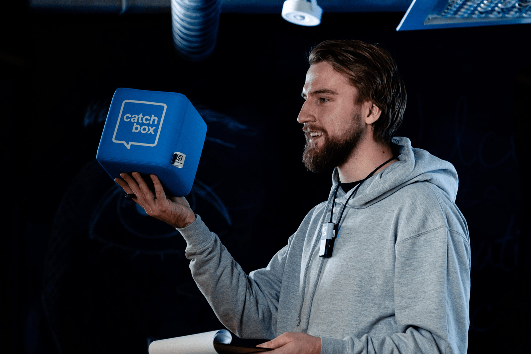 Presenter holding a blue Catchbox Cube microphone, wearing a Catchbox Clip microphone, in a dimly lit room with a focused expression, enhancing conference accessibility with clear audio for live captions.