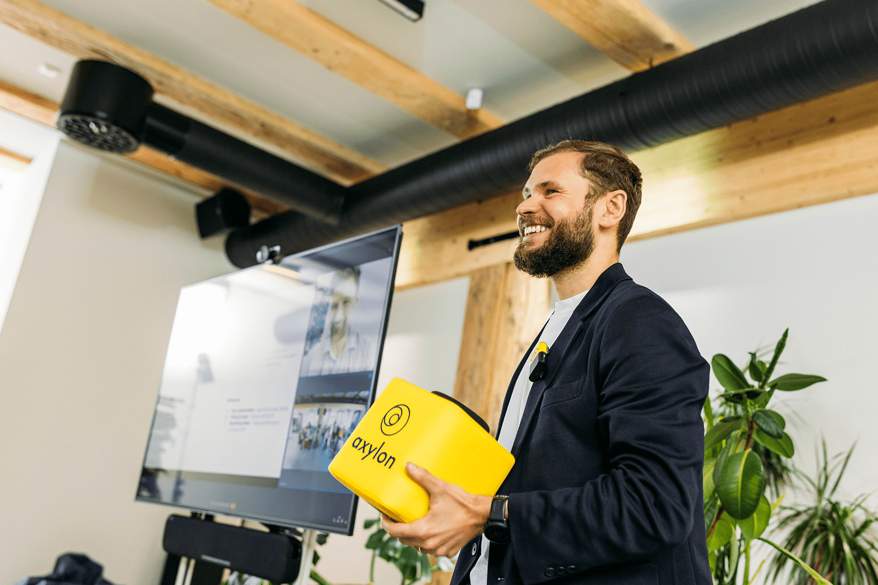 A smiling presenter holding a yellow Catchbox Cube microphone during a hybrid meeting, with a screen in the background displaying remote participants.